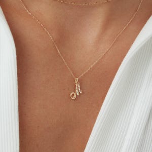 May include: Gold-toned necklace with two delicate chains. The lower chain features a pendant in the shape of the letter "M" embellished with small, sparkling stones. The necklace is worn against a white shirt.