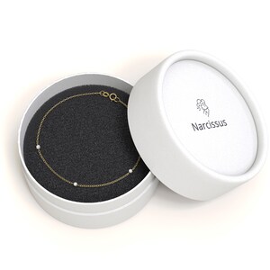 May include: A delicate gold chain bracelet with three small white stones, presented in a white box with the brand name "Narcissus" printed on the lid.