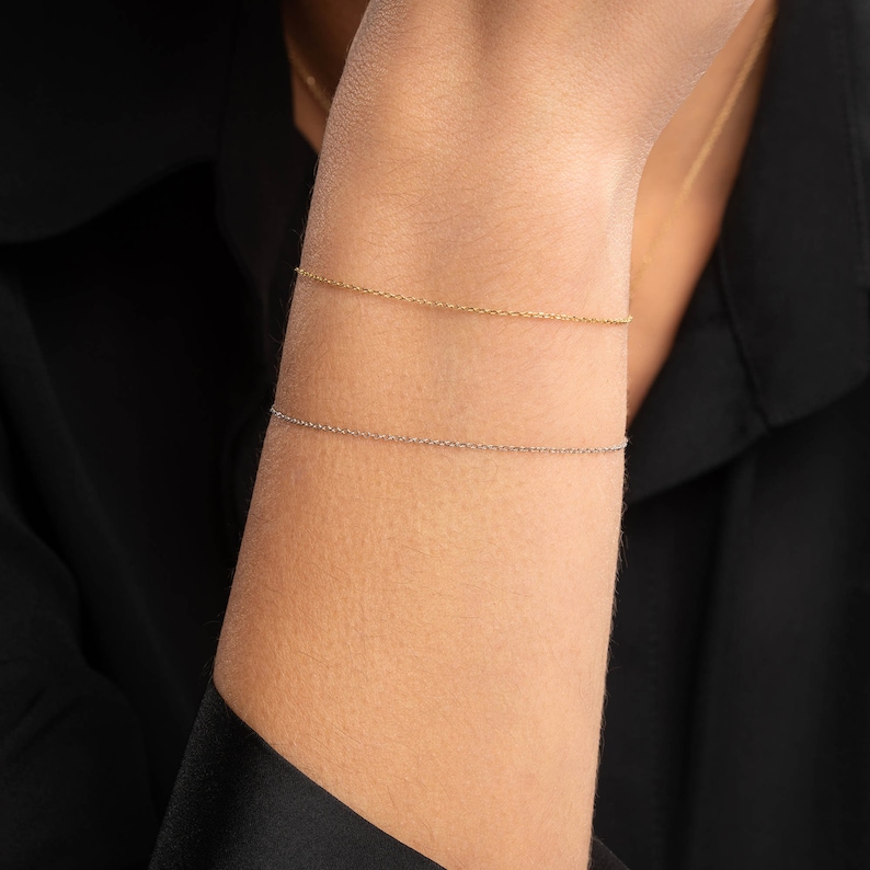 May include: Two delicate chain bracelets, one gold and one silver, worn on a person's wrist.