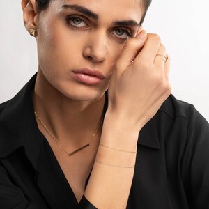 May include: A woman wearing a black blazer with a gold bar necklace and two gold chain bracelets. She is wearing a gold triangle earring and a gold ring on her finger.
