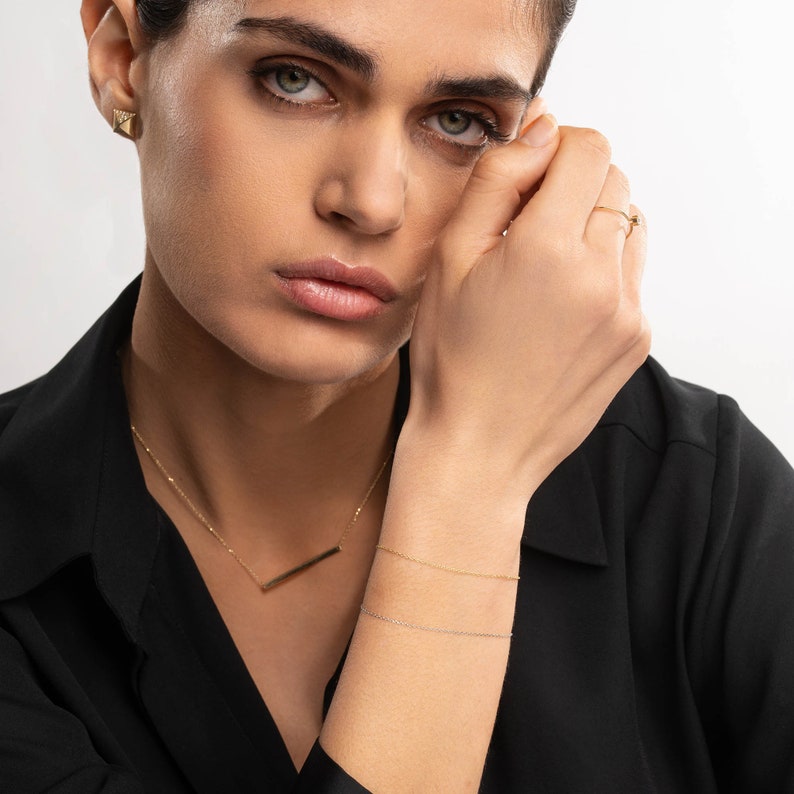 May include: A woman wearing a black blazer with a gold bar necklace and two gold chain bracelets. She is wearing a gold triangle earring and a gold ring on her finger.