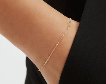 May include: A delicate gold bracelet with a chain-link design. The bracelet is worn on a person's wrist, with a black garment in the background. The bracelet features small, evenly spaced beads and links.