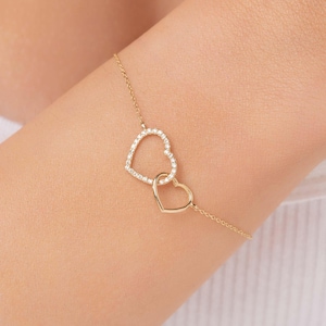 May include: A gold bracelet featuring two interlocking heart charms. The hearts are set with small, clear stones.