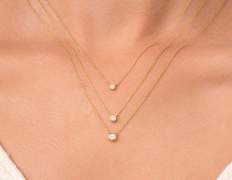May include: Three gold necklaces with small round clear stones hanging from them. The necklaces are layered on a person's neck.