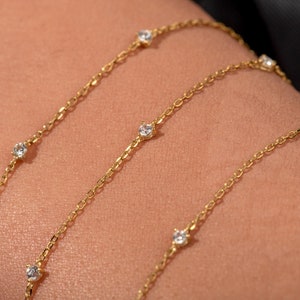 May include: A gold chain necklace with small, clear gemstones. The necklace is layered on a person's arm.