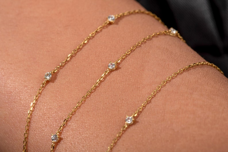 May include: A gold chain necklace with small, clear gemstones. The necklace is layered on a person's arm.