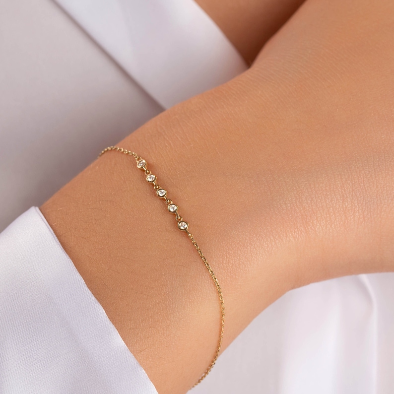 Pu&ograve; includere: Un delicato braccialetto d'oro con una serie di piccoli diamanti scintillanti. Il braccialetto &egrave; una catena sottile con sei diamanti rotondi distanziati uniformemente. Il gioiello &egrave; esposto su un polso, contro una camicia bianca.