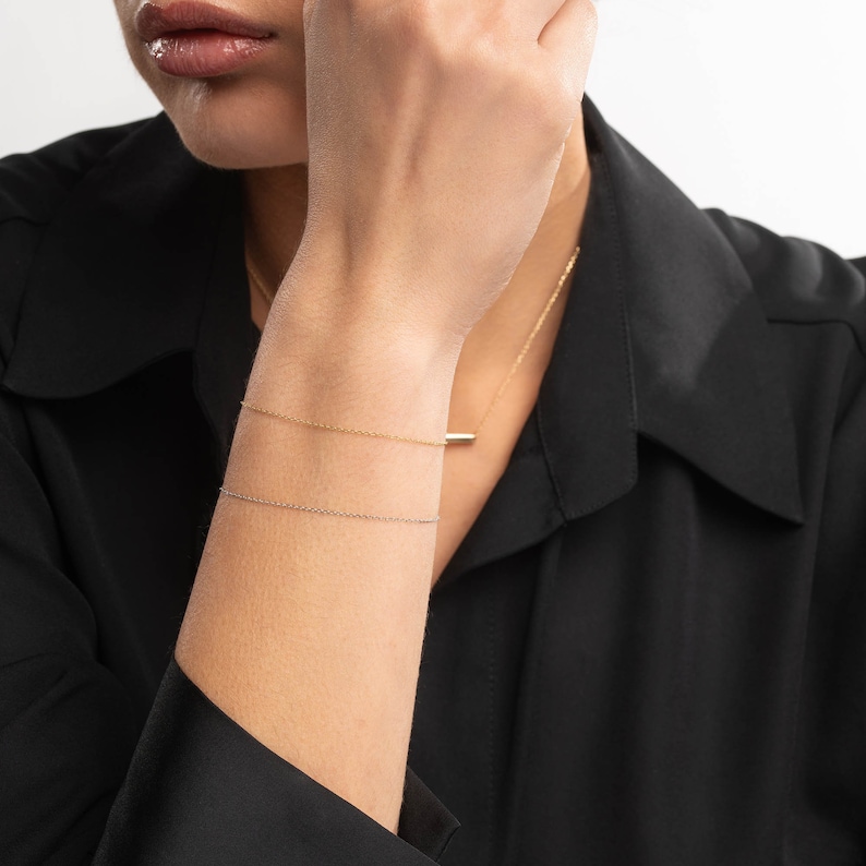 May include: Two delicate gold chain bracelets on a person's wrist. The bracelets are layered, with a thinner chain on top of a thicker chain.