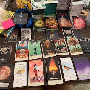 May include: A spread of tarot and oracle cards on a wooden table. The cards feature various illustrations, including a cauldron, a mouse, a tree, and a bird. The cards are arranged in rows and columns, with some cards overlapping. The cards are surrounded by other items, including books, a pink sticky note, and a pair of headphones.