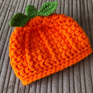 May include: An orange crochet hat with green leaf detailing, resembling a carrot. The hat is made from textured yarn. The hat is suitable for babies and children. The hat's circumference is approximately 40 cm.