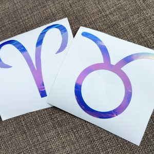 May include: Two iridescent vinyl stickers featuring the astrological signs of Aries and Taurus. The stickers are on a white background.
