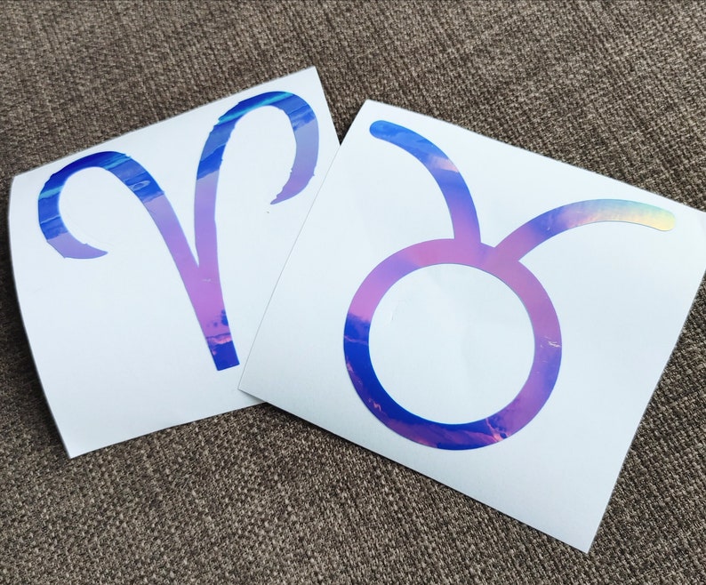 May include: Two iridescent vinyl stickers featuring the astrological signs of Aries and Taurus. The stickers are on a white background.