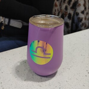 May include: A purple stainless steel tumbler with a holographic sticker of the Libra zodiac sign.