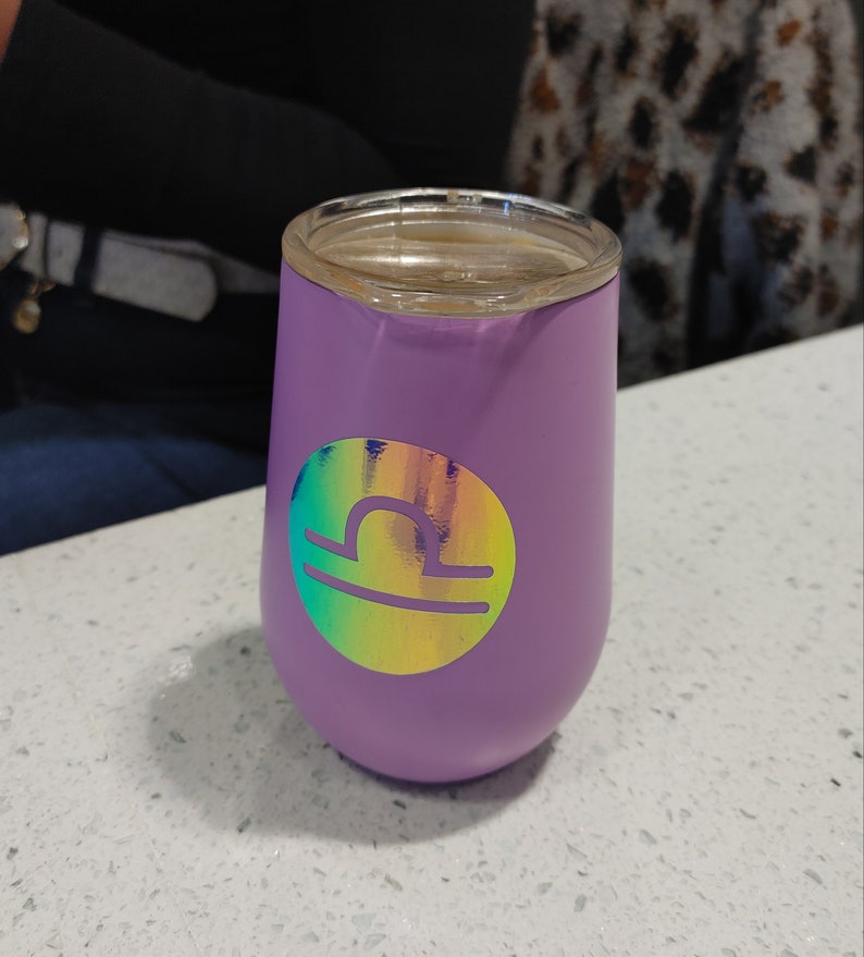 May include: A purple stainless steel tumbler with a holographic sticker of the Libra zodiac sign.