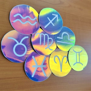 May include: A set of nine iridescent holographic stickers featuring the astrological signs. The stickers are round and have a white outline. The signs are: Aquarius, Taurus, Gemini, Cancer, Leo, Virgo, Libra, Sagittarius, and Capricorn.