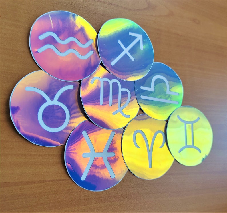 May include: A set of nine iridescent holographic stickers featuring the astrological signs. The stickers are round and have a white outline. The signs are: Aquarius, Taurus, Gemini, Cancer, Leo, Virgo, Libra, Sagittarius, and Capricorn.
