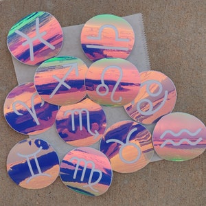 May include: Twelve iridescent holographic stickers featuring the astrological signs. Each sticker has a white outline and a white zodiac symbol on a pink, blue, and purple background.