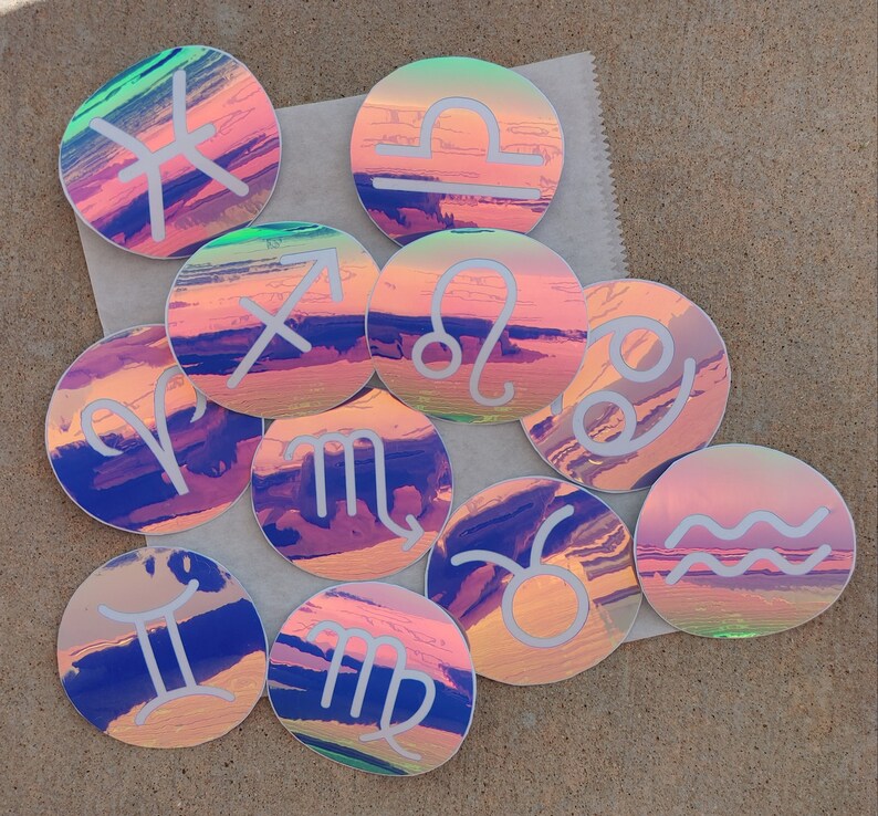May include: Twelve iridescent holographic stickers featuring the astrological signs. Each sticker has a white outline and a white zodiac symbol on a pink, blue, and purple background.