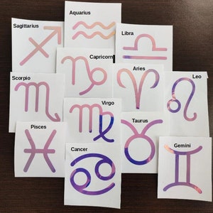 May include: Twelve white cards with pink and purple holographic zodiac signs. Each card features a different zodiac sign with its name printed above it. The signs include: Sagittarius, Aquarius, Libra, Capricorn, Aries, Leo, Scorpio, Virgo, Taurus, Gemini, Pisces, and Cancer.