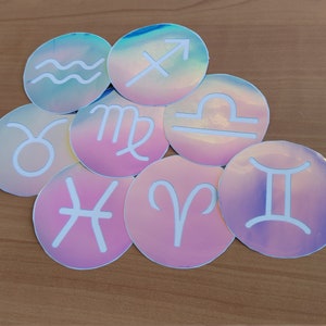 May include: Set of 10 iridescent holographic stickers featuring the astrological zodiac signs. Each sticker is round and features a white zodiac symbol on a pink or blue background.