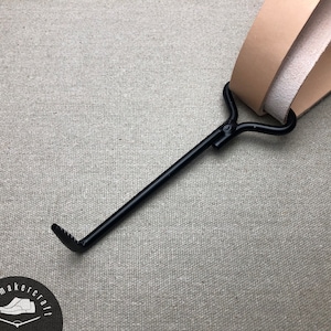 Puede incluir: Un calzador de metal negro con un mango curvo y un extremo dentado. El calzador se combina con correas de cuero color beige. El logotipo de shoemaker's craft es visible en la esquina inferior izquierda.