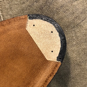 Puede incluir: Primer plano de una suela de zapato de cuero. La suela es marr&oacute;n con un ribete negro y una inserci&oacute;n de color beige. Se ven tres peque&ntilde;os agujeros en la inserci&oacute;n. El fondo es una tela texturizada de color neutro.