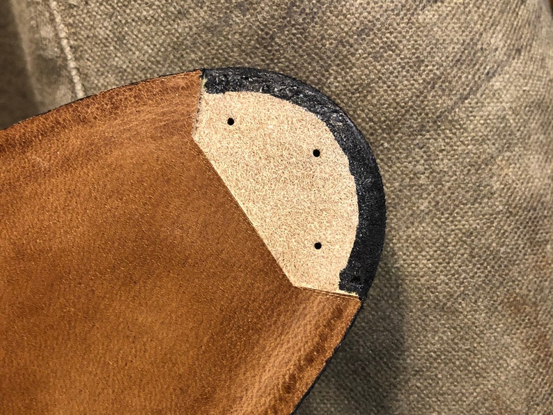 Puede incluir: Primer plano de una suela de zapato de cuero. La suela es marr&oacute;n con un ribete negro y una inserci&oacute;n de color beige. Se ven tres peque&ntilde;os agujeros en la inserci&oacute;n. El fondo es una tela texturizada de color neutro.