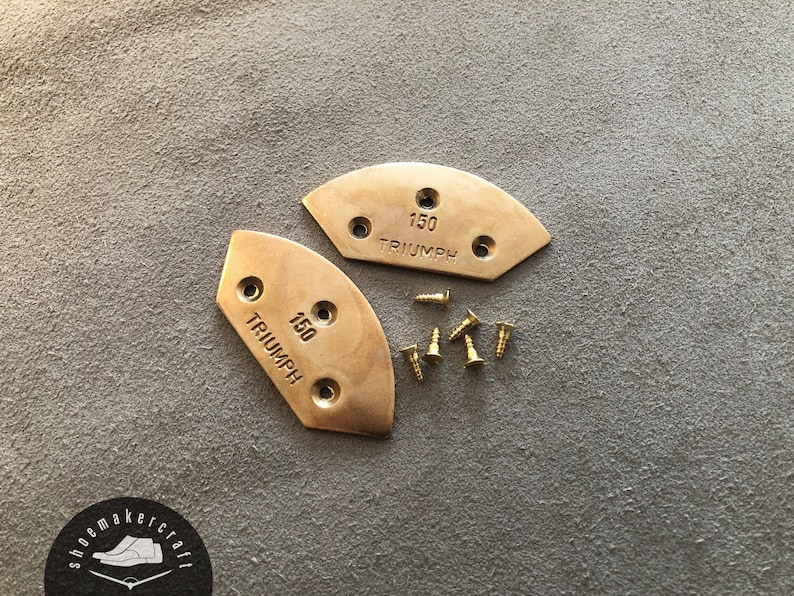 Puede incluir: Dos protectores de suela de zapato Triumph 150 de lat&oacute;n con tornillos de montaje. Los protectores son semicirculares con tres agujeros y el texto "150 TRIUMPH". Los tornillos son peque&ntilde;os y dorados.