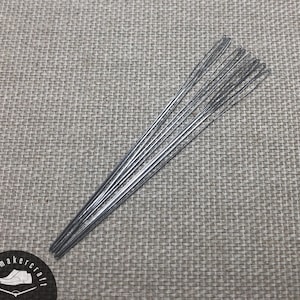 Puede incluir: Un conjunto de agujas plateadas de zapatero dispuestas sobre una superficie texturizada de color gris claro. Las agujas son finas y tienen un pequeño ojo para enhebrar. La imagen también incluye un logotipo circular con el texto "shoemakercraft".