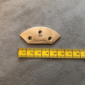 Puede incluir: Una placa semicircular de color lat&oacute;n con tres agujeros y el texto "150 TRIUMPH" se muestra. Una regla amarilla con marcas negras se coloca debajo. La placa est&aacute; sobre una superficie gris.
