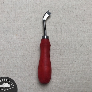 May include: A leatherworking tool with a curved, silver blade and a red wooden handle. The tool is designed for precision cutting and shaping of leather materials. The handle is approximately 4 inches long.