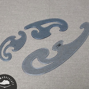 Puede incluir: Tres reglas de curva francesa claras y curvas de varios tamaños. Estas herramientas de dibujo están hechas de plástico transparente y se utilizan para dibujar curvas suaves en diseño y dibujo técnico. Las reglas están dispuestas sobre una superficie gris.