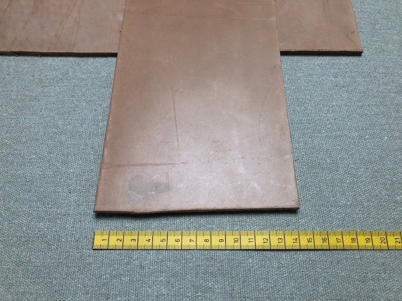 K&ouml;nnte beinhalten: Nahaufnahme von mehreren St&uuml;cken braunen Leders, m&ouml;glicherweise zum Basteln. Ein gelbes Lineal mit schwarzen Markierungen liegt am unteren Rand und zeigt Messungen bis zu 53 cm an. Die Lederst&uuml;cke liegen auf einer grauen Stoffoberfl&auml;che.