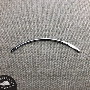 May include: A curved, silver shoemaker's needle, resting on a textured grey surface. The needle has a sharp, pointed end and a blunt, rectangular end. The needle is slightly curved.