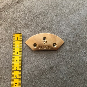 Puede incluir: Una placa met&aacute;lica de color lat&oacute;n, semicircular, con el texto "150 TRIUMPH" y tres agujeros. Una regla amarilla est&aacute; colocada junto a la placa, mostrando medidas. La placa es probablemente una pieza o un emblema.