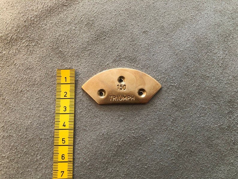 Puede incluir: Una placa met&aacute;lica de color lat&oacute;n, semicircular, con el texto "150 TRIUMPH" y tres agujeros. Una regla amarilla est&aacute; colocada junto a la placa, mostrando medidas. La placa es probablemente una pieza o un emblema.