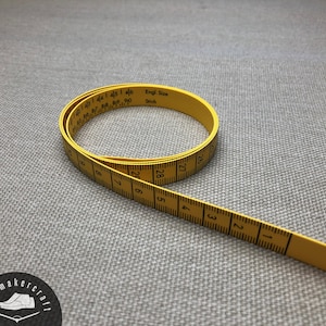 May include: A yellow measuring tape with black markings and numbers, coiled on a grey surface. The tape is marked in centimetres and has the text "Engl. Size Stitch". Suitable for sewing and craft projects.