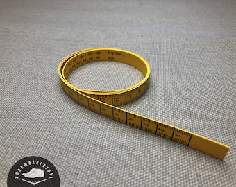 Könnte beinhalten: Ein gelbes Maßband mit schwarzen Markierungen und Zahlen, auf einer grauen Oberfläche aufgerollt. Das Band ist in Zentimetern markiert und trägt den Text "Engl. Size Stitch". Ideal für Näh- und Bastelprojekte.