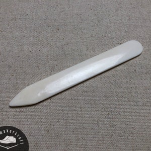 May include: A bone folder tool, a craft essential, is displayed on a neutral-toned fabric. The tool is a pale ivory color, with a smooth, rounded end and a pointed tip. It is used for scoring, folding, and burnishing paper and other materials.