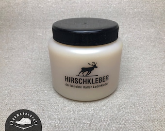 Könnte beinhalten: Ein weißes Glas Hirschkleber Lederkleber mit schwarzem Deckel. Auf dem Glas ist der Markenname und ein Hirschlogo abgebildet. Darunter steht "der beliebte Haller Lederkleber". Für Lederarbeiten.