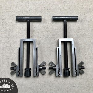 May include: Two metal shoe making clamps with a T-shaped handle, threaded rod, and wing nuts. The clamps are silver with black threaded rods and wing nuts. The background is a neutral beige fabric.