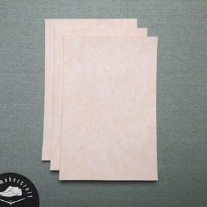 Puede incluir: Tres hojas rectangulares de cuero de color beige claro están apiladas sobre una superficie gris. El cuero tiene un aspecto moteado. Las hojas están dispuestas verticalmente, con los bordes superiores alineados. El logotipo de Shoemaker Craft es visible en la esquina inferior izquierda.