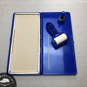 May include: Open blue ink pad case with a beige pad, a small roller, a metal pin, and a blue and black stamp. The case is open, revealing the tools inside. The items are for crafting.