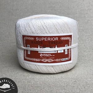 Puede incluir: Un carrete de hilo de ramio superior blanquecino, 450g, de Tosco Co. Ltd, Tokio, Japón. La etiqueta es de color marrón rojizo con texto blanco y un borde decorativo. El hilo está bien enrollado y asegurado con una cuerda blanca.