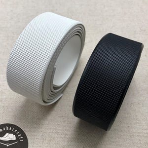 May include: Two rolls of textured material, one white and one black, are displayed. The material has a repeating pattern of small, raised squares. The rolls are unrolled, revealing the inner side. The image is taken on a neutral-colored surface.