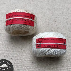 Puede incluir: Dos bobinas de hilo blanquecino con etiquetas rojas. Las etiquetas dicen "SPAGO PER CALZOLAI" y "MARCA CERVO". El hilo está bien enrollado y las etiquetas están envueltas alrededor del centro. El fondo es una tela beige claro.