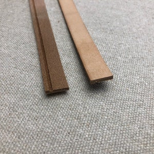 K&ouml;nnte beinhalten: Zwei Streifen braunes Leder werden vor einem neutralen Hintergrund gezeigt. Ein Streifen ist ein dunkleres, strukturiertes Braun, w&auml;hrend der andere ein helleres, glattes Tan ist. Die Streifen sind rechteckig und scheinen f&uuml;r Bastelarbeiten oder Lederarbeiten bestimmt zu sein.