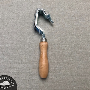 Pode incluir: Ferramenta de sapateiro com cabo de madeira e cabeça metálica curva com pequenos dentes. A ferramenta, provavelmente para fabricação de calçados, está sobre um fundo cinza.