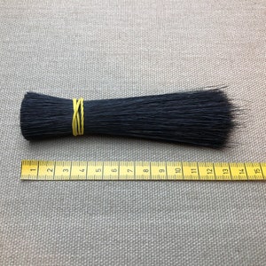 Może przedstawiać: Pęczek czarnych włosia, zabezpieczony ż&oacute;łtą gumką, leży obok ż&oacute;łtej miarki. Włosie ma około 18 cm długości. Tło stanowi neutralna szara tkanina.