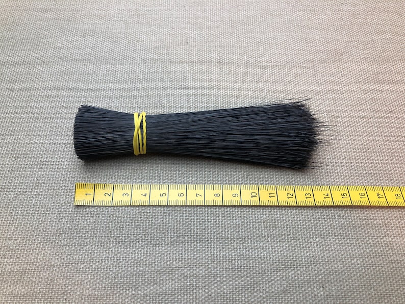 Może przedstawiać: Pęczek czarnych włosia, zabezpieczony ż&oacute;łtą gumką, leży obok ż&oacute;łtej miarki. Włosie ma około 18 cm długości. Tło stanowi neutralna szara tkanina.
